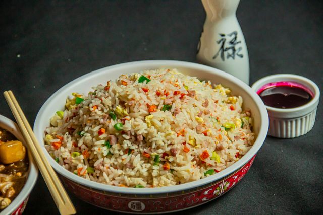 Tazón de arroz frito con zanahorias, guisantes y huevo, con palillos, salsa y una botella de sake sobre un fondo oscuro.
