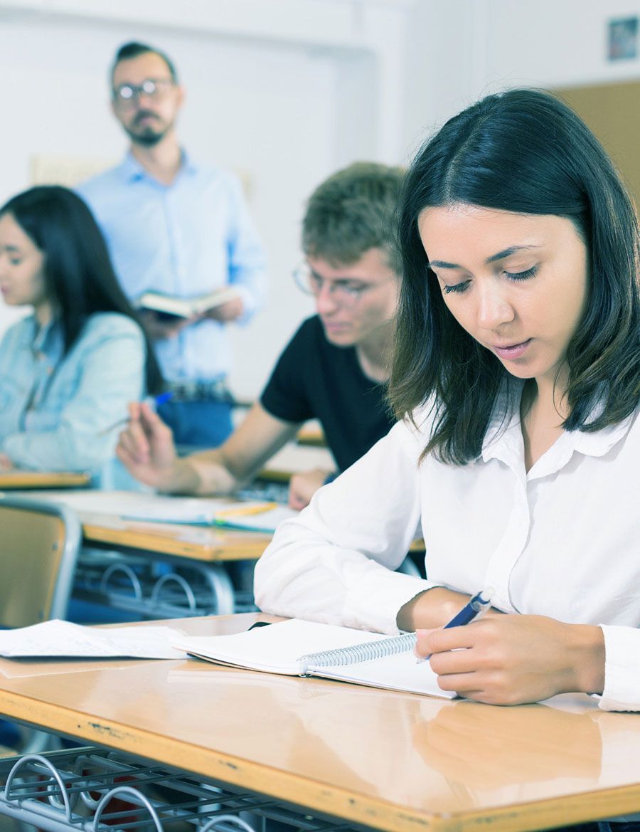 Una mujer está sentada en un escritorio en un aula escribiendo en un cuaderno.
