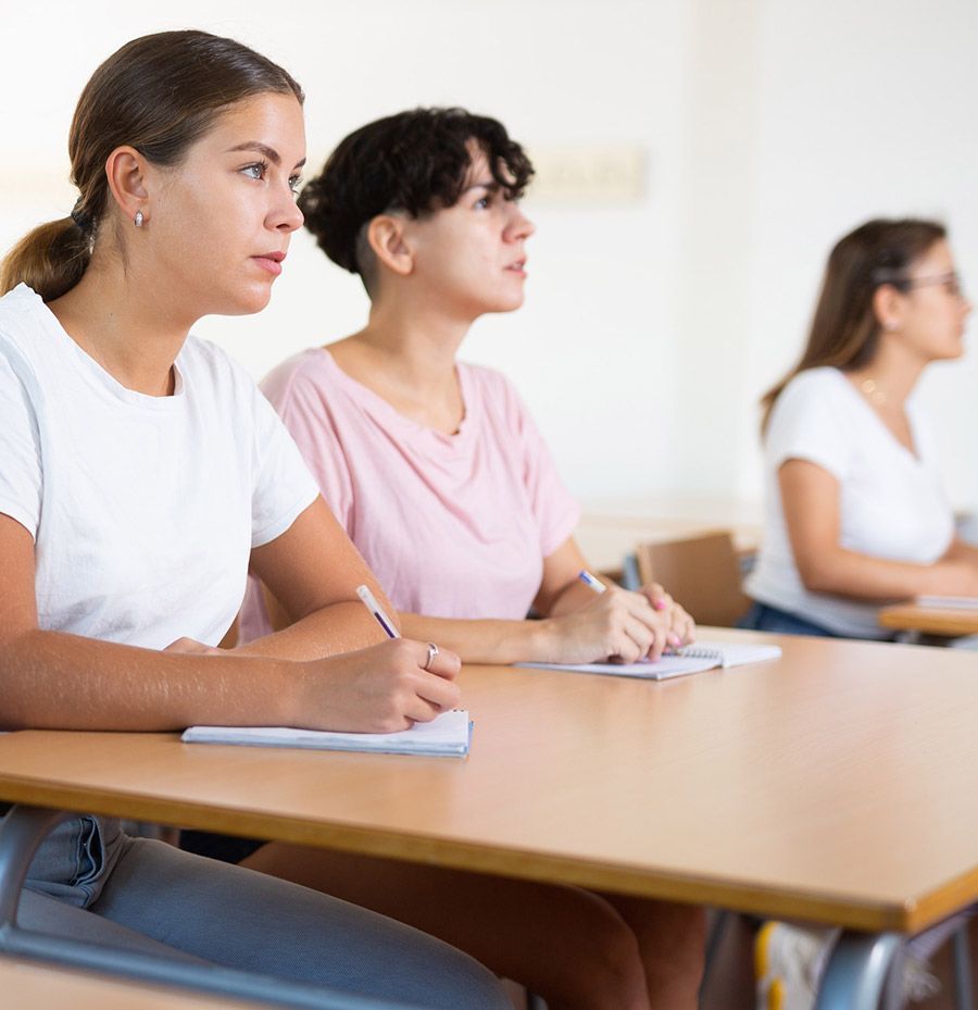 Tres mujeres se sientan en escritorios en un aula escribiendo en cuadernos.