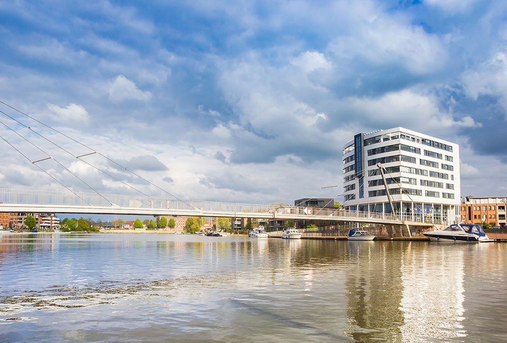 Uferszene mit Brücke, modernem Gebäude, Booten und bewölktem Himmel.