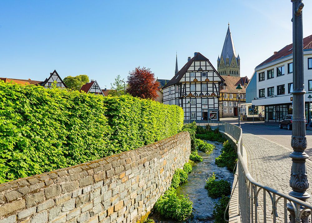 Steinmauer und Hecke an einem Bach in einer deutschen Stadt; im Hintergrund Fachwerkhäuser und Kirchturm.