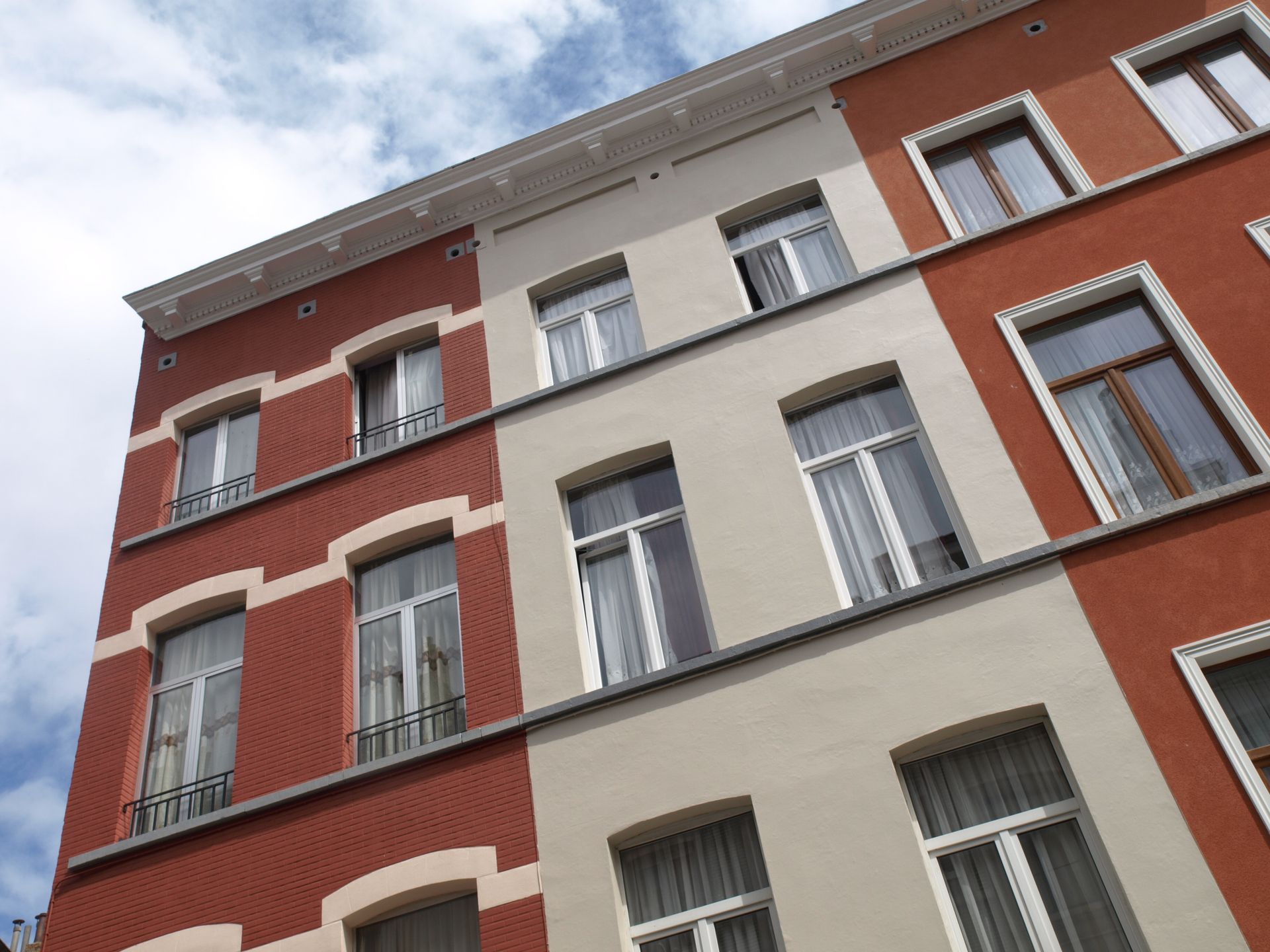 Fachada de edificio de apartamentos de color rojo y crema con múltiples ventanas, vista en ángulo contra un cielo azul.