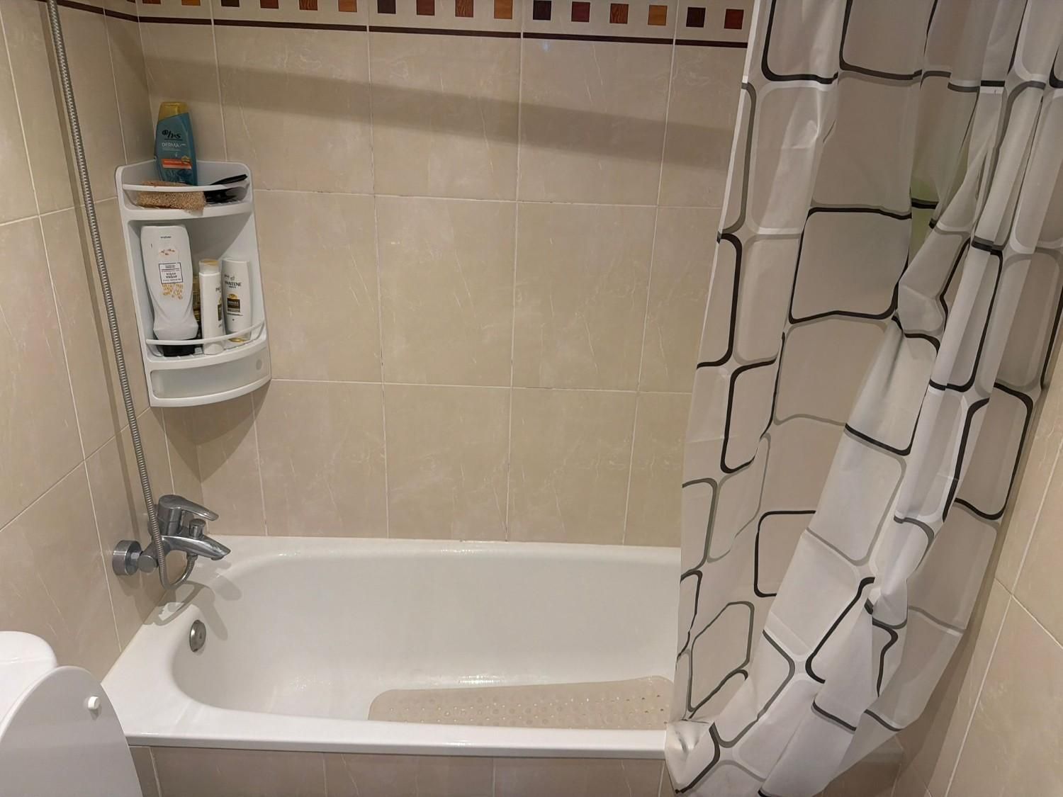 Baño pequeño de color beige con bañera, cortina de ducha y estante de pared para artículos de aseo.