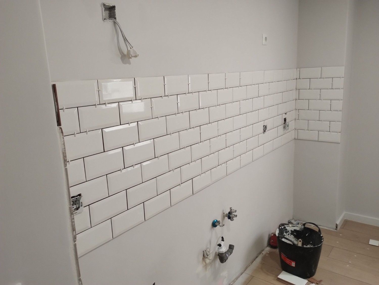 Pared parcialmente alicatada en una cocina en reforma con tomas de agua a la vista y una bolsa de herramientas en el suelo.