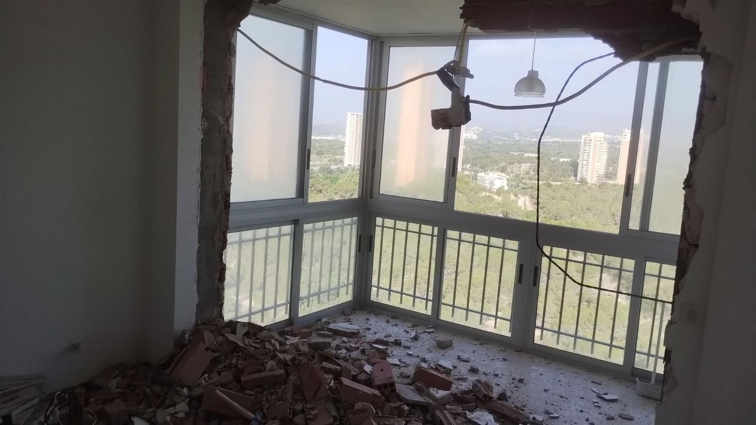 Habitación dañada con escombros en el suelo y una ventana de balcón de esquina rota con vistas a la ciudad.