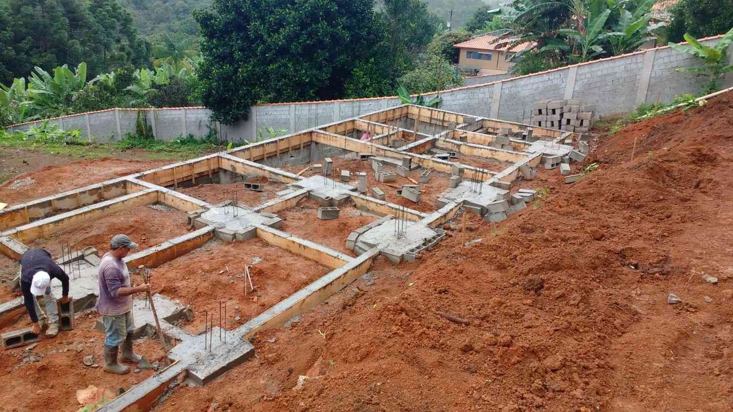 Obra en construcción con cimientos, trabajadores y tierra roja en una ladera.