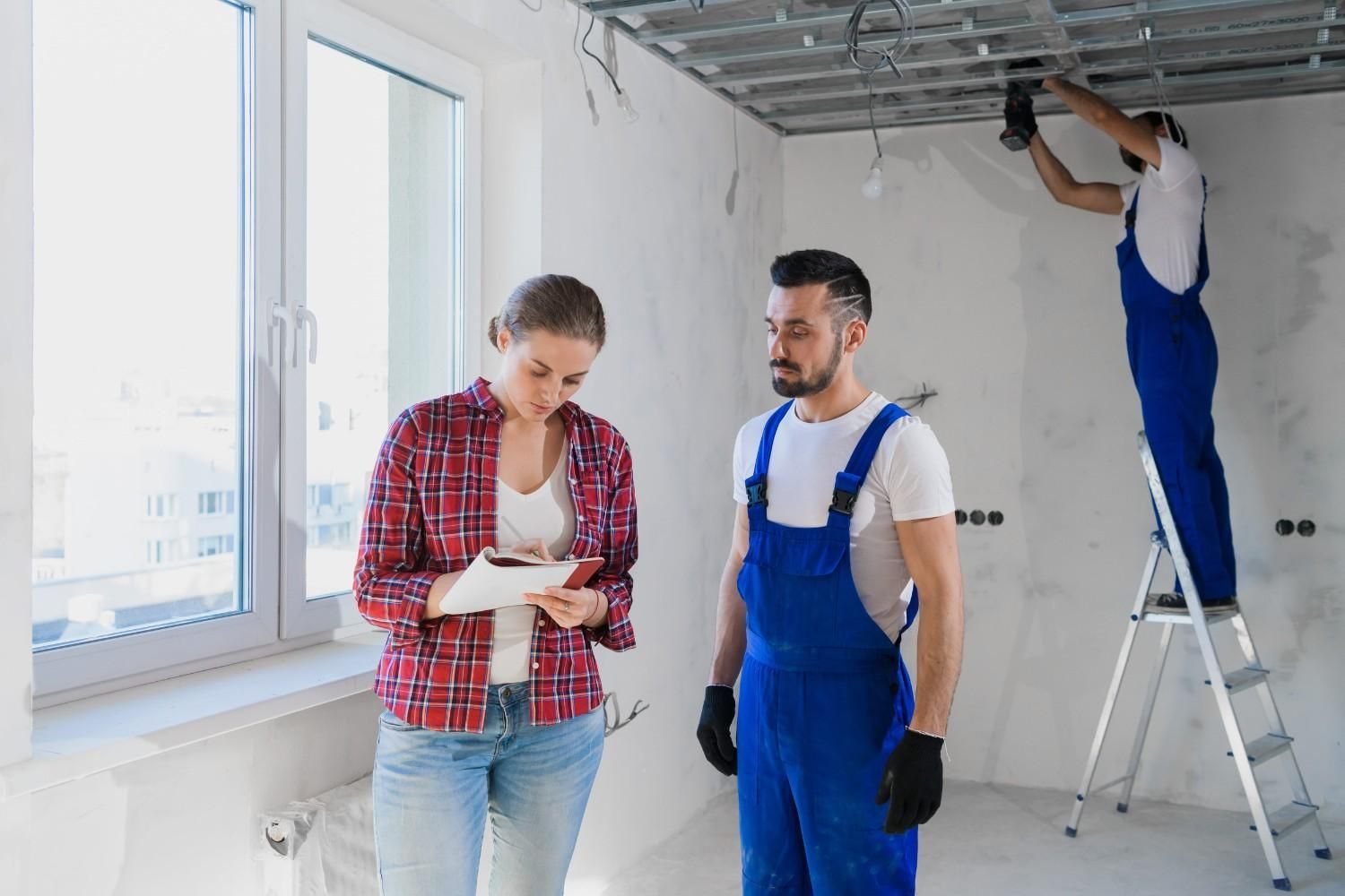 Dos trabajadores con monos azules inspeccionan la reforma de una habitación mientras otro pinta el techo.