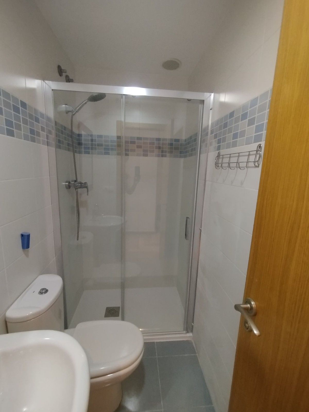 Baño pequeño con inodoro, lavabo y ducha con mampara de cristal, azulejos blancos y borde decorativo azul.