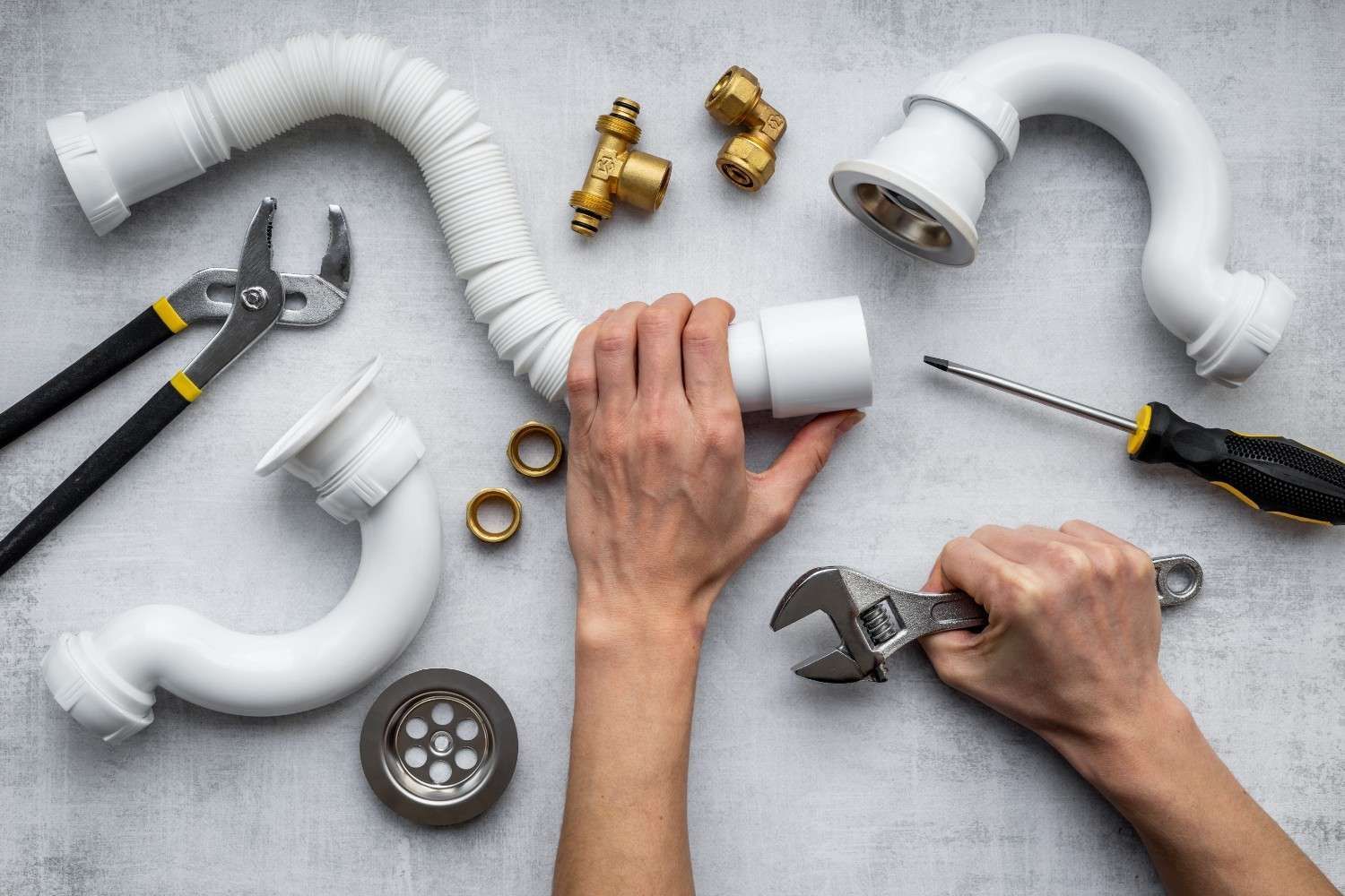 Manos ensamblando tuberías de PVC blancas con herramientas y accesorios de latón sobre una superficie gris.