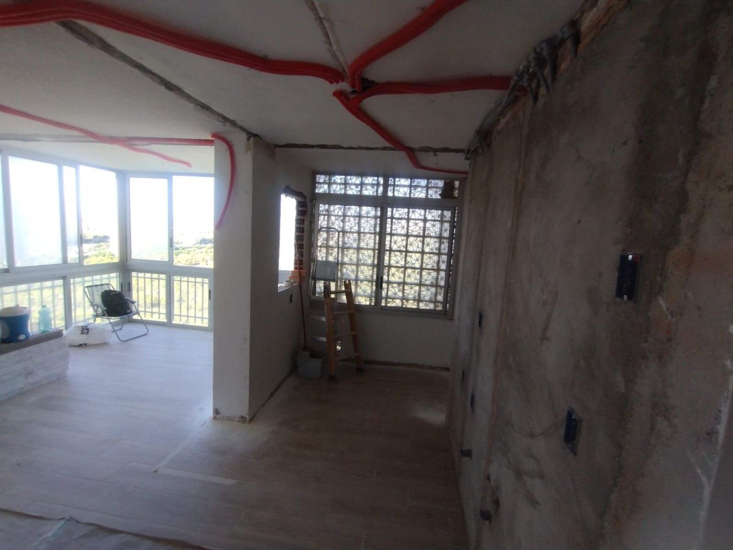 Habitación vacía sin terminar, con cableado a la vista, paredes grises y vistas a un balcón a través de grandes ventanales.