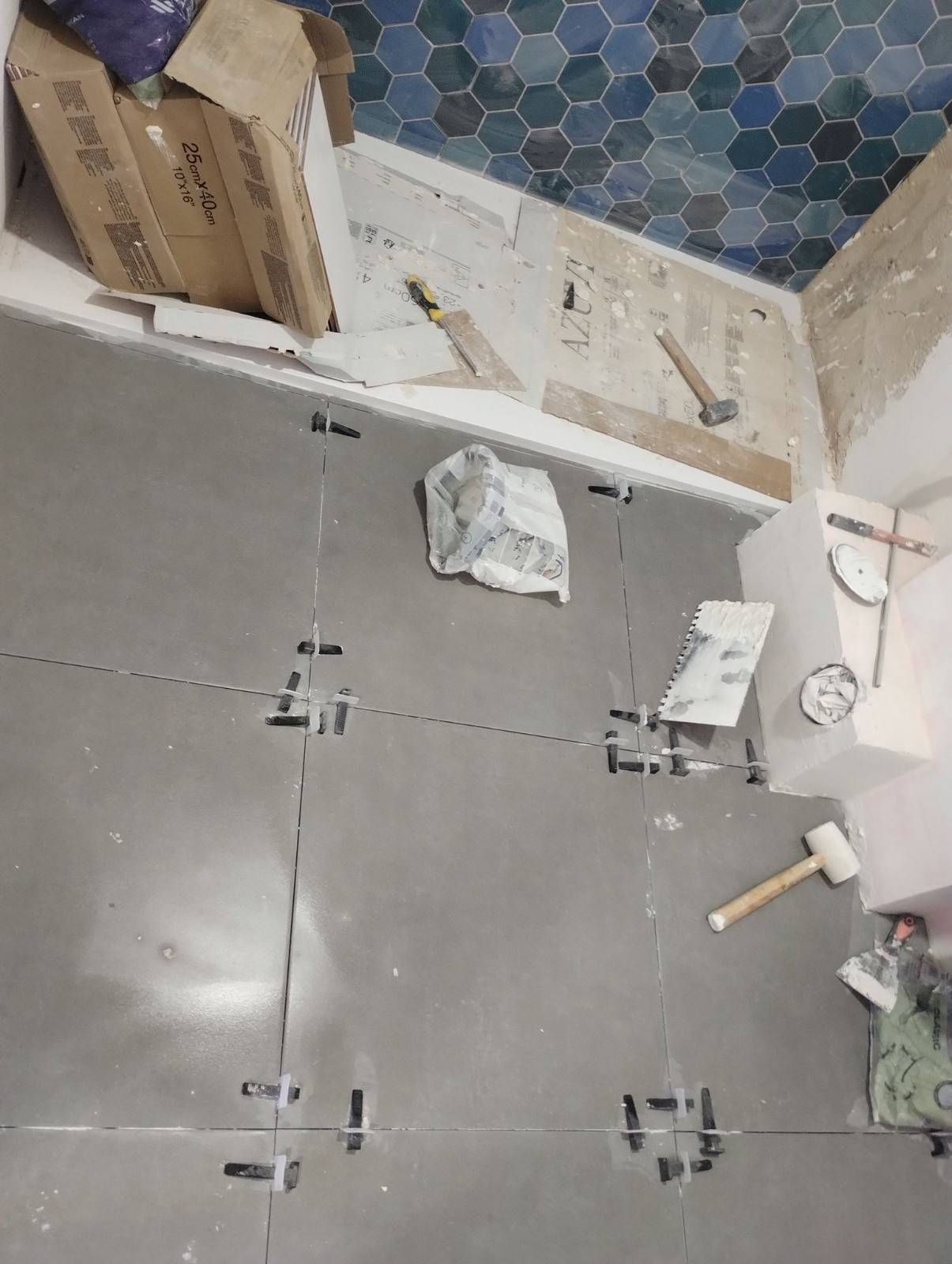 Piso del baño en construcción con azulejos grises, mosaico en la pared y herramientas y materiales dispersos.