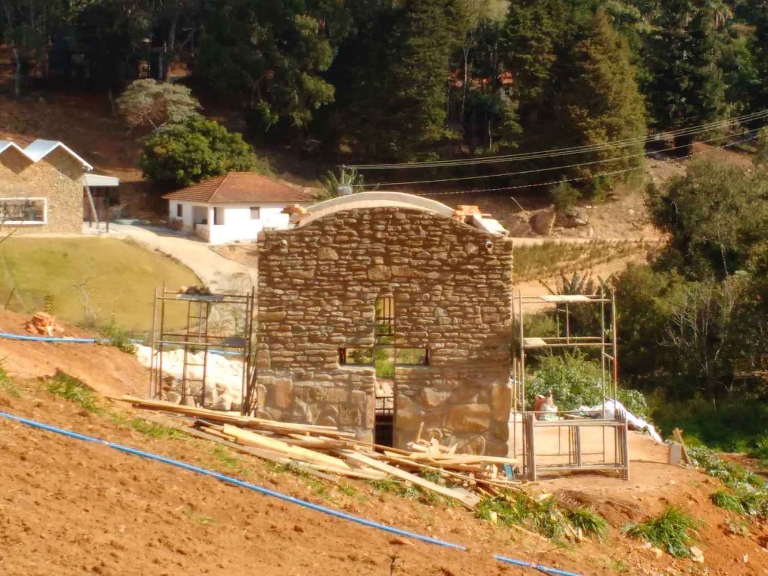 Edificio de piedra en construcción en una ladera inclinada con andamios y árboles al fondo.