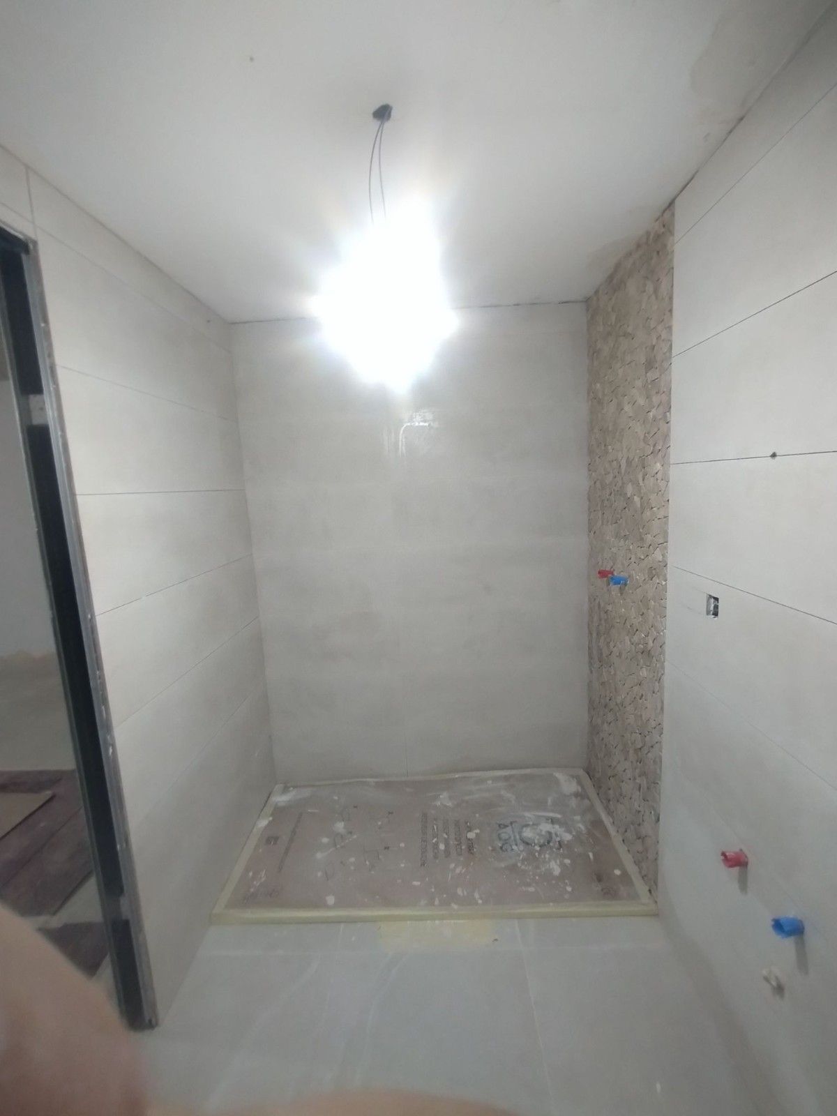Pequeña zona de ducha de baño alicatada en construcción, con paredes sin revestir y lámpara colgante.