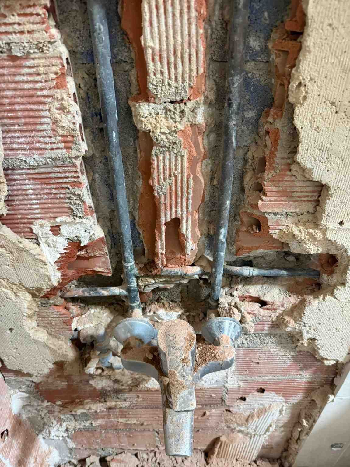 Tuberías y válvulas de fontanería expuestas en una pared de ladrillo dañada con parches de yeso y hormigón rugosos.