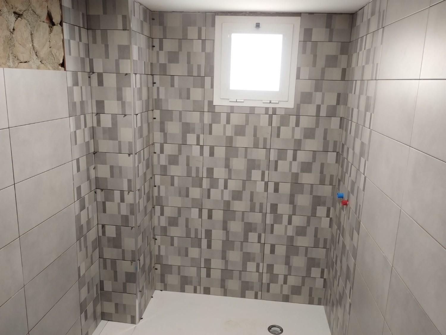 Baño pequeño con ducha alicatada, pared decorativa con mosaico gris y una pequeña ventana sobre la pared del fondo.