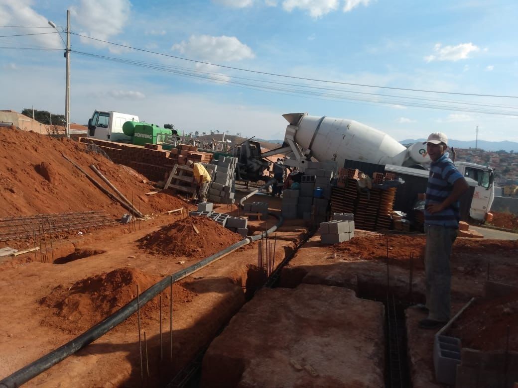 Obra en construcción con zanja de tierra roja, camión hormigonera y trabajador bajo un cielo azul.