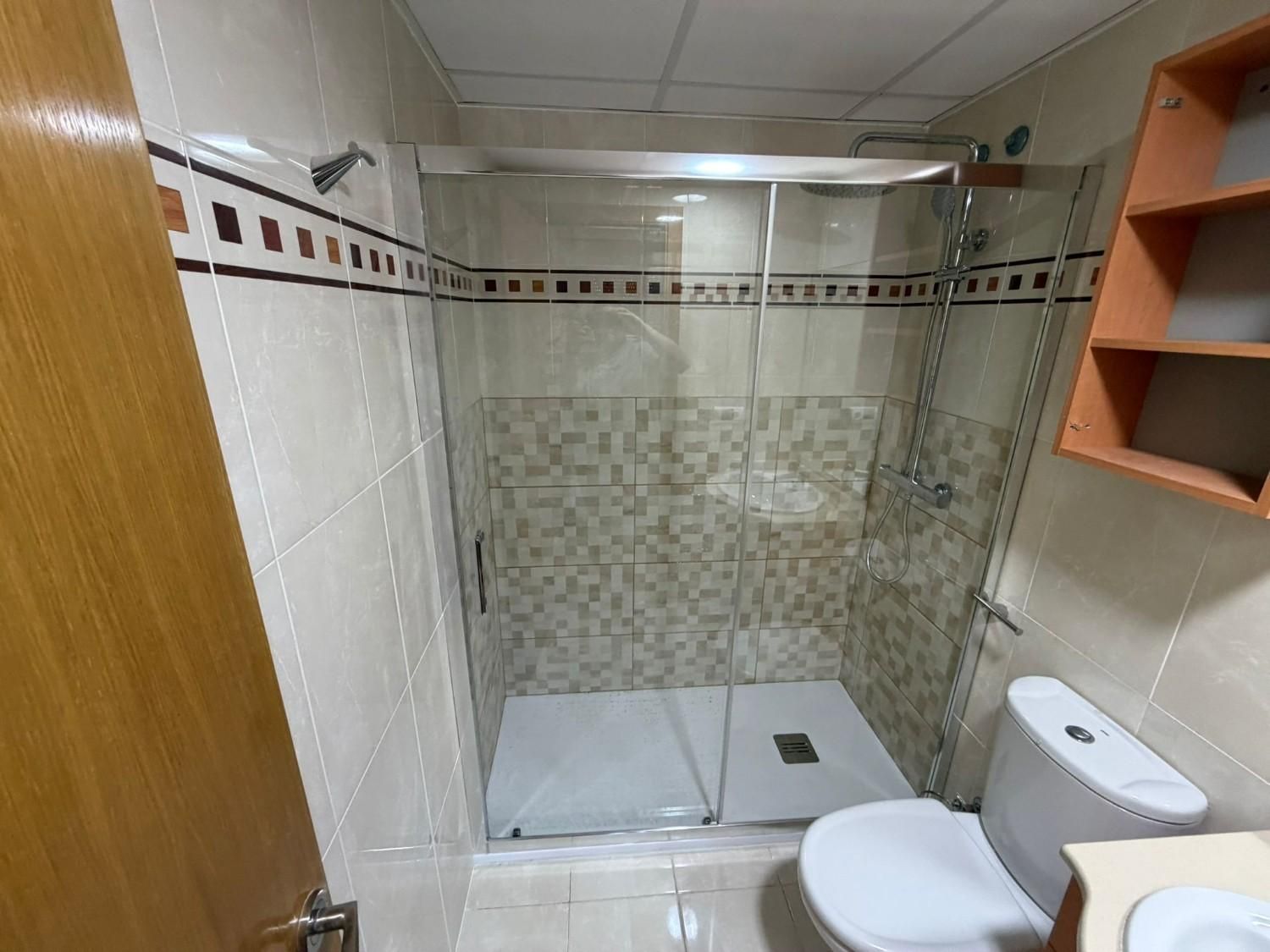 Baño pequeño con azulejos, cabina de ducha de cristal, inodoro blanco y estantes en la pared.