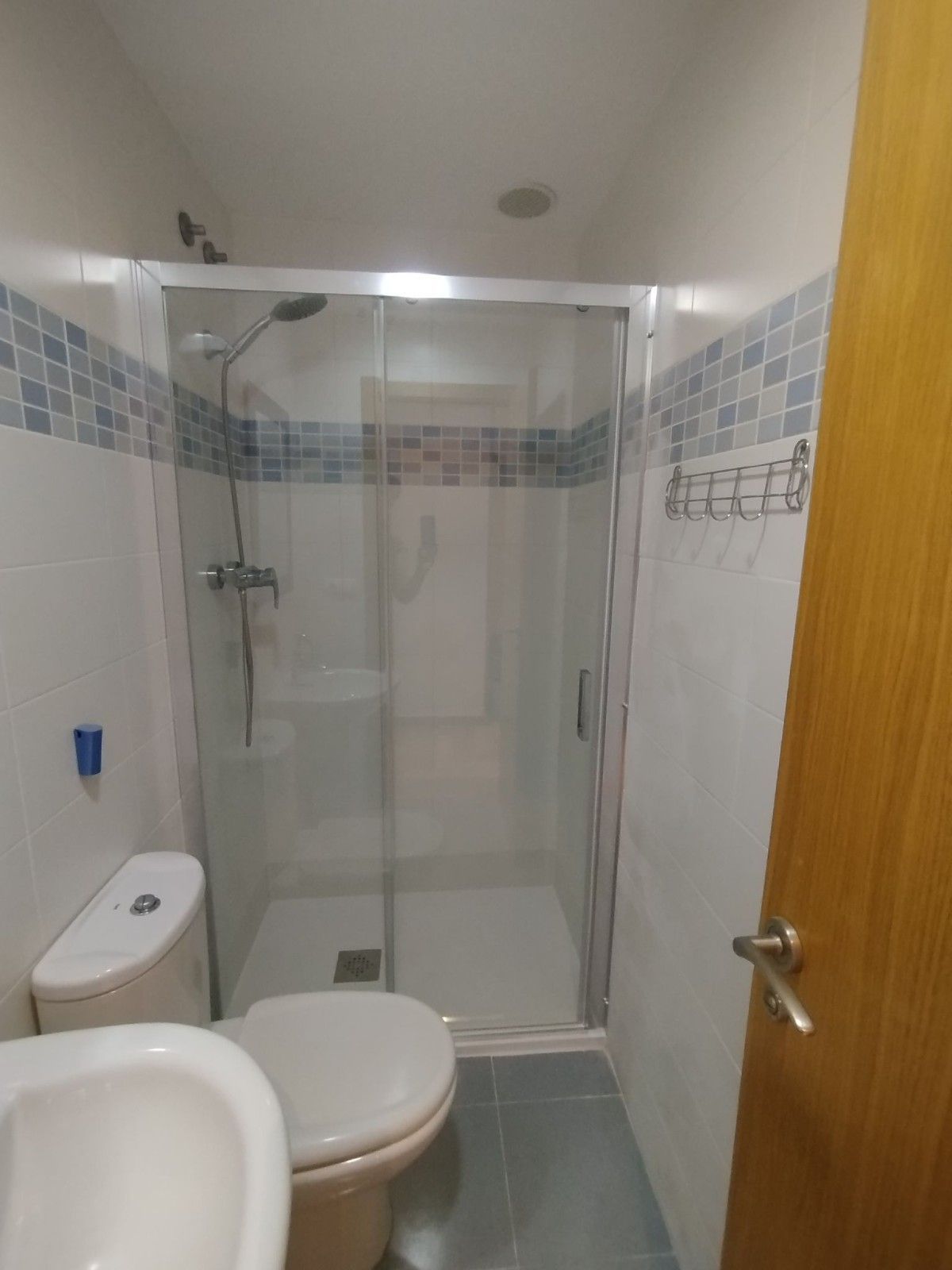 Baño pequeño con inodoro, lavabo y cabina de ducha de cristal; azulejos blancos con una franja decorativa azul.