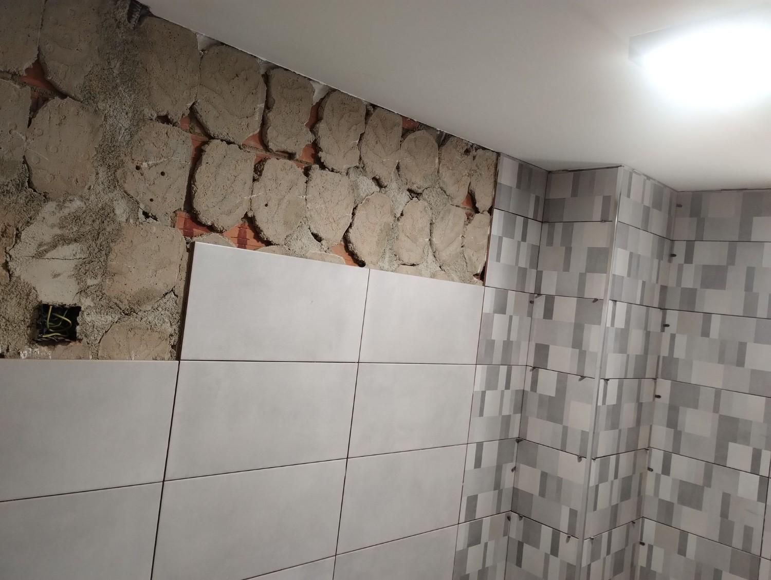 Pared del baño en proceso de renovación con piedra sin acabado, azulejos blancos y azulejos grises con estampado.