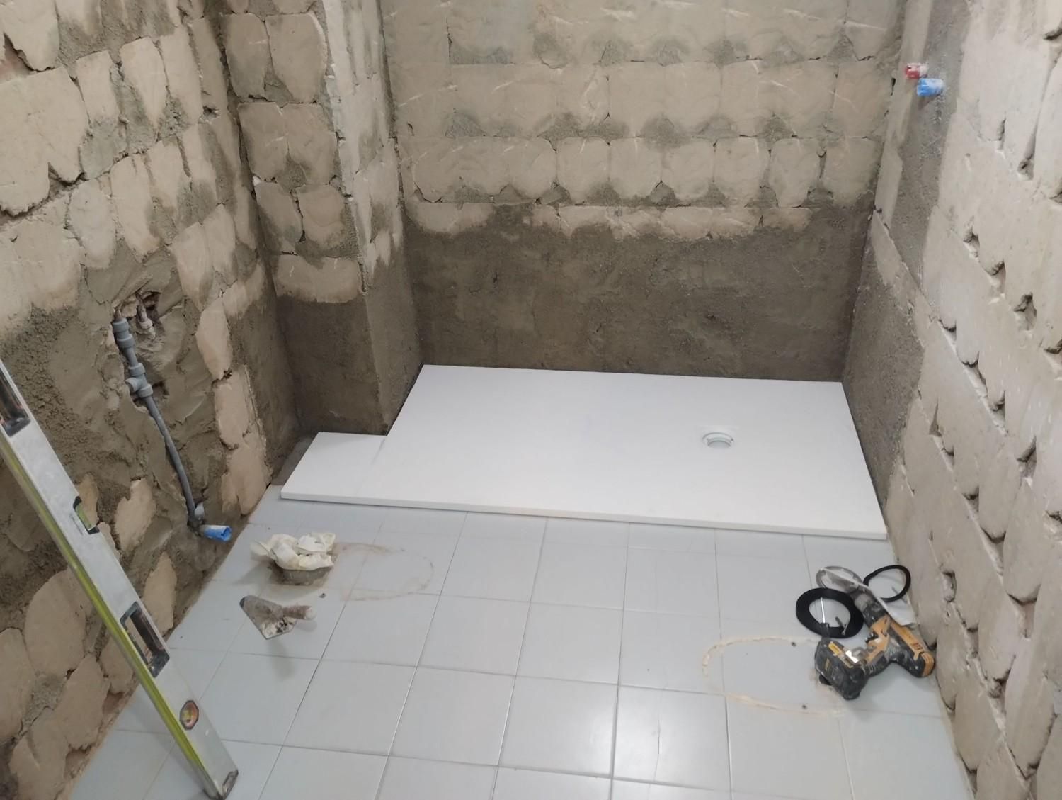 Baño pequeño sin terminar con paredes y suelo alicatados, plato de ducha, tuberías a la vista y herramientas de construcción.