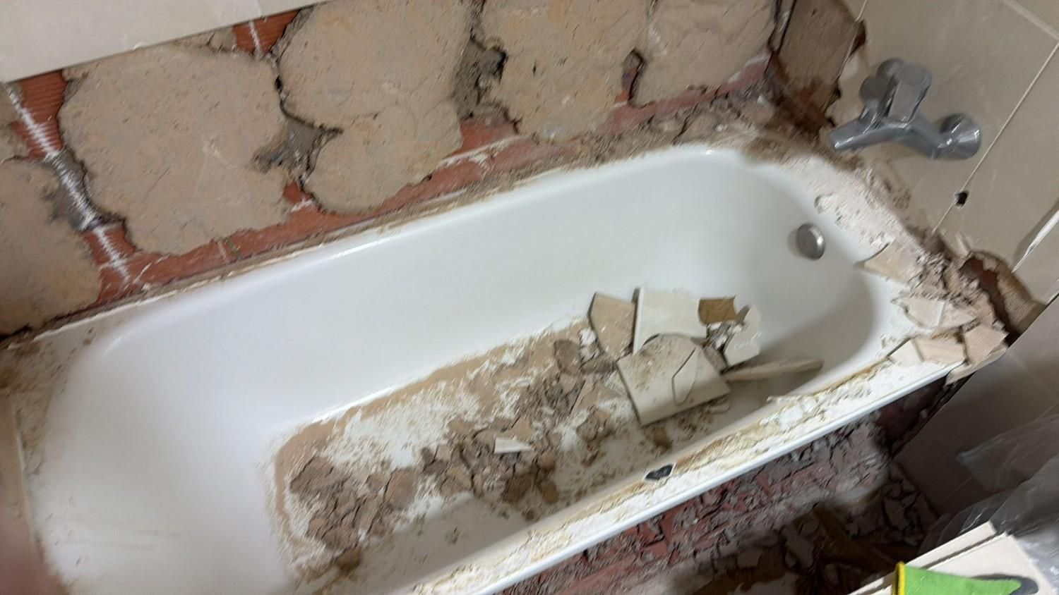 Bañera blanca dañada con azulejos de pared rotos y escombros en un baño descuidado.