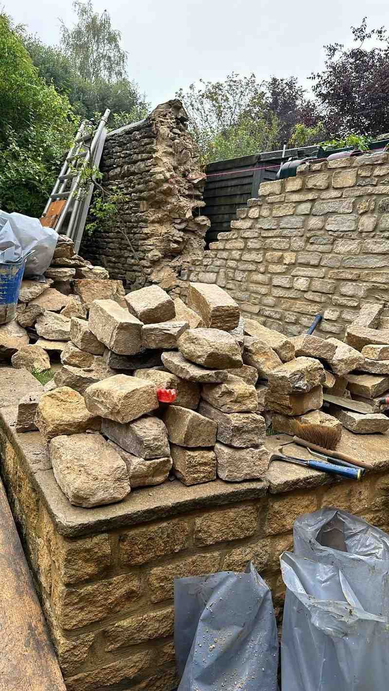Muro de piedra derrumbado con escombros y restos esparcidos al aire libre, con árboles al fondo.