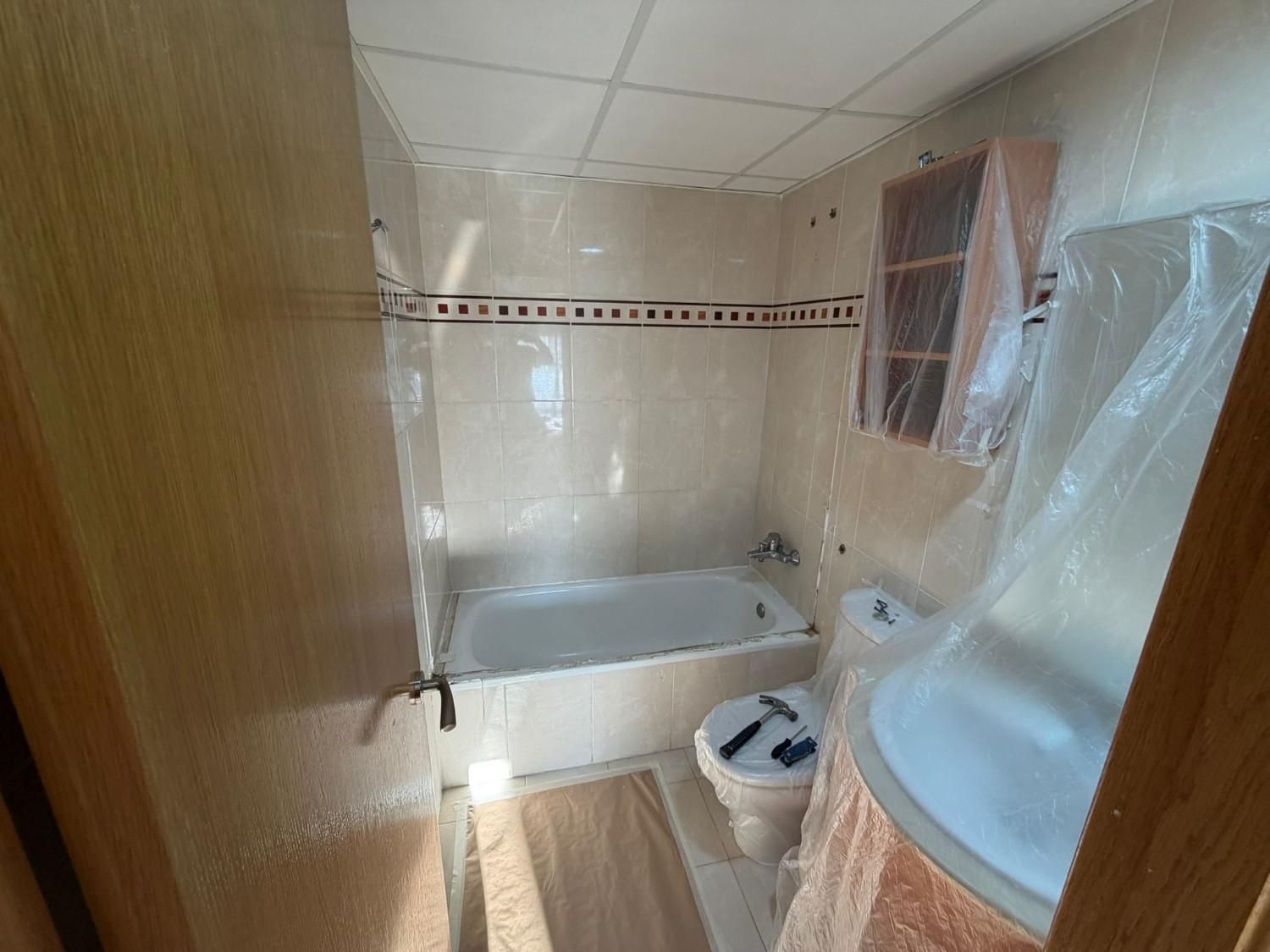 Baño pequeño con bañera, inodoro, lavabo y puerta con paneles de madera cubierta con plástico protector.