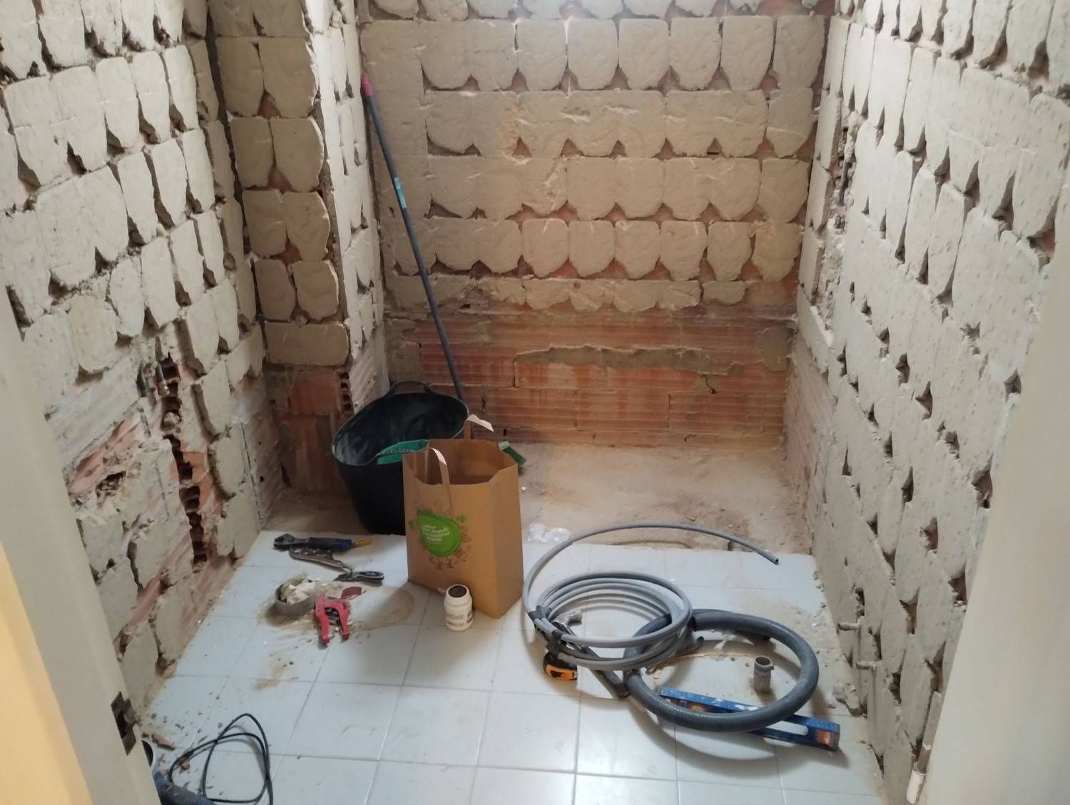Una pequeña habitación con azulejos, herramientas de construcción, cables, un cubo y una caja de cartón en el suelo.