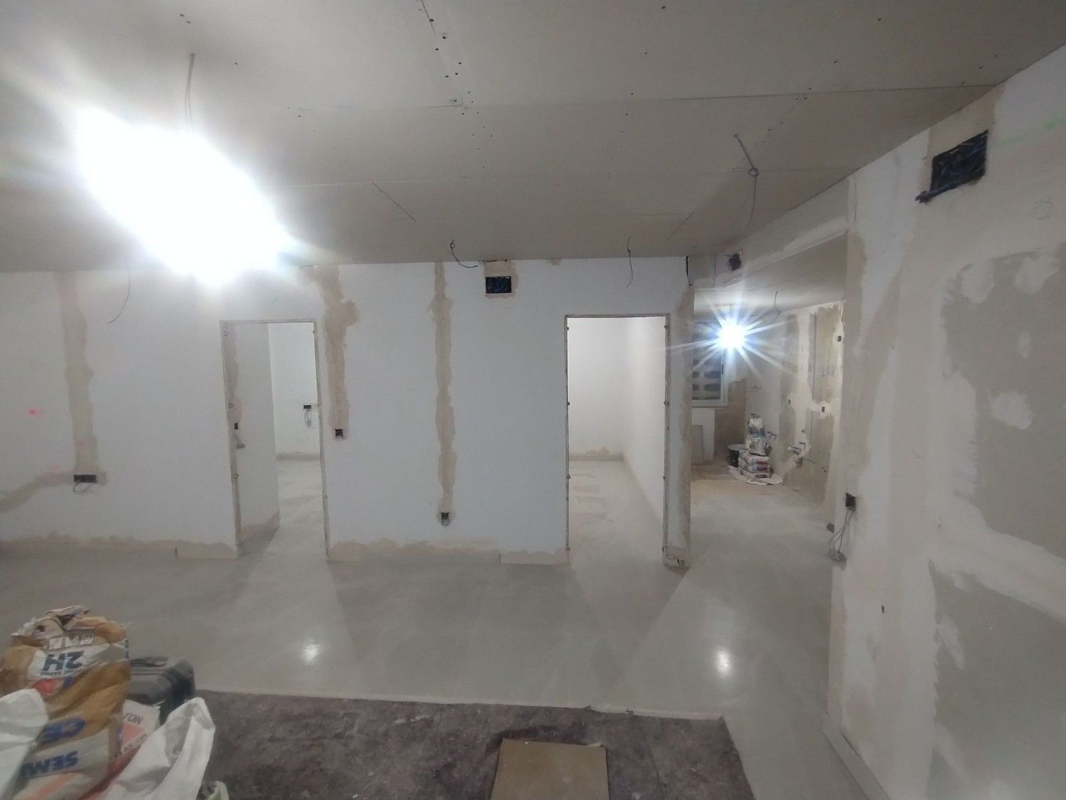 Habitación vacía sin terminar con paredes blancas, luces brillantes en el techo y materiales de construcción en el suelo.