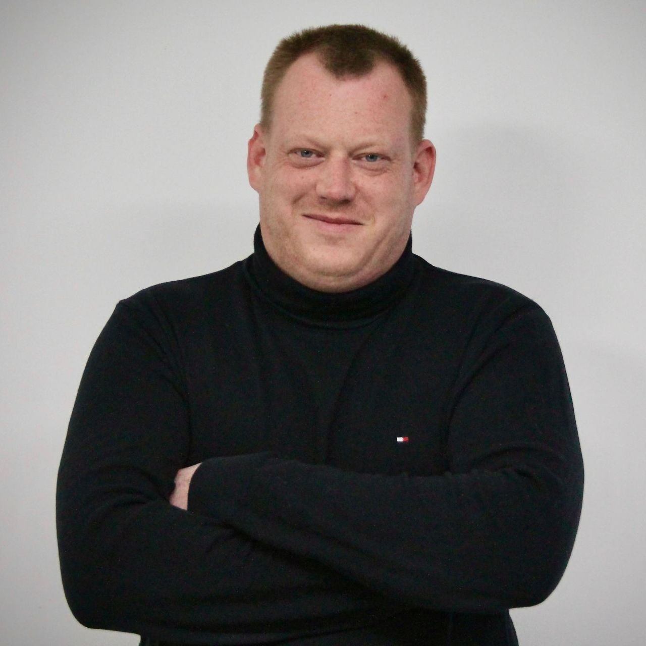 ein Mann trägt einen schwarzen Rollkragenpullover und lächelt mit verschränkten Armen