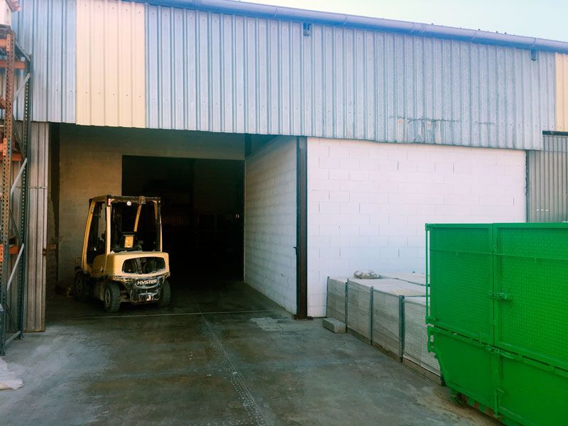 Una carretilla elevadora amarilla está estacionada afuera de un almacén al lado de un contenedor de basura verde.