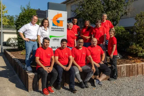 Eine Gruppe von Menschen in roten Hemden posiert im Freien neben einem Firmenschild in einer sonnigen, gepflegten Landschaft.