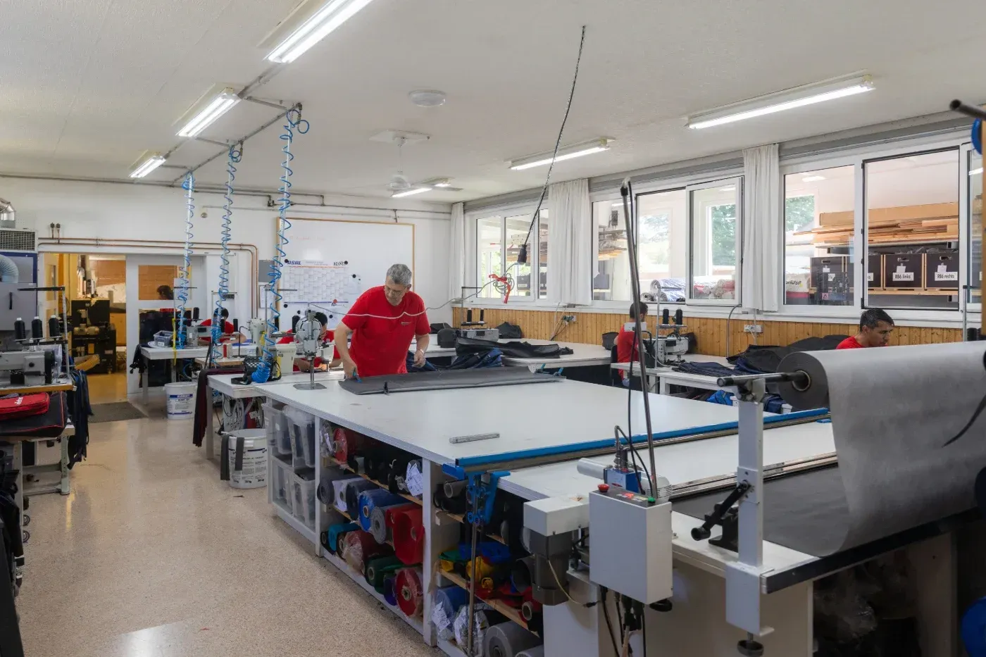 Helle Nähwerkstatt mit großen Arbeitstischen, Stoffrollen und einer Arbeiterin in einem roten Hemd.