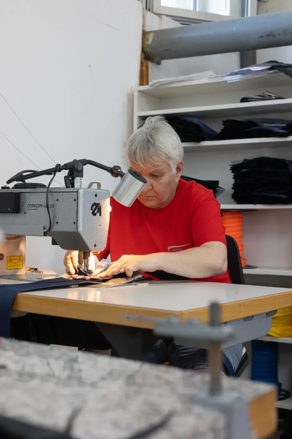 Eine Person in einem roten Hemd, die in einer Werkstatt an einer Nähmaschine arbeitet und sich auf Stoffarbeiten konzentriert.