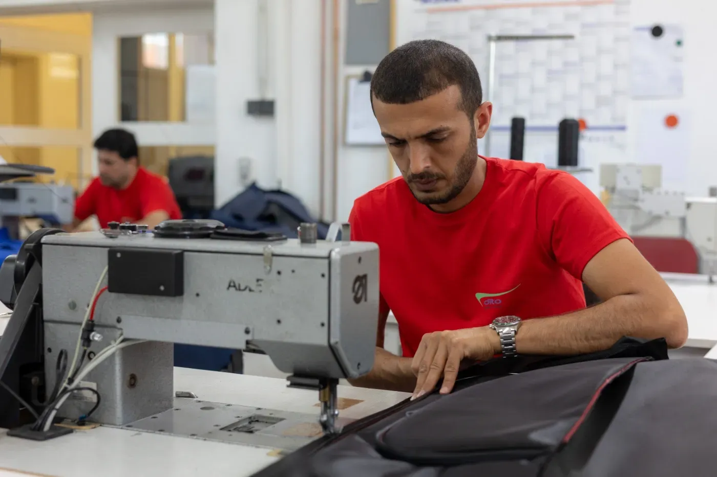 Ein Schneider im roten Hemd arbeitet in einer Werkstatt mit einer Nähmaschine an schwarzem Stoff.