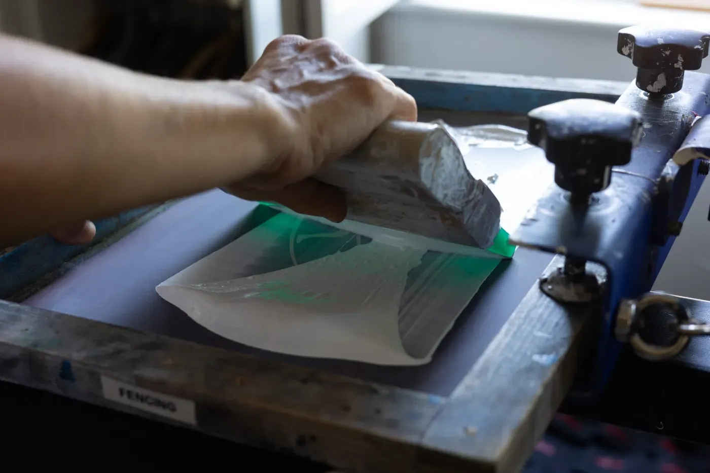Mit einer Siebdruckpresse ein grünes Design von Hand auf ein weißes T-Shirt drucken.