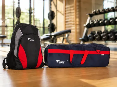 Schwarze und rote Sporttasche und Reisetasche auf einem Holztisch in einem Fitnessraum, im Hintergrund Hanteln.