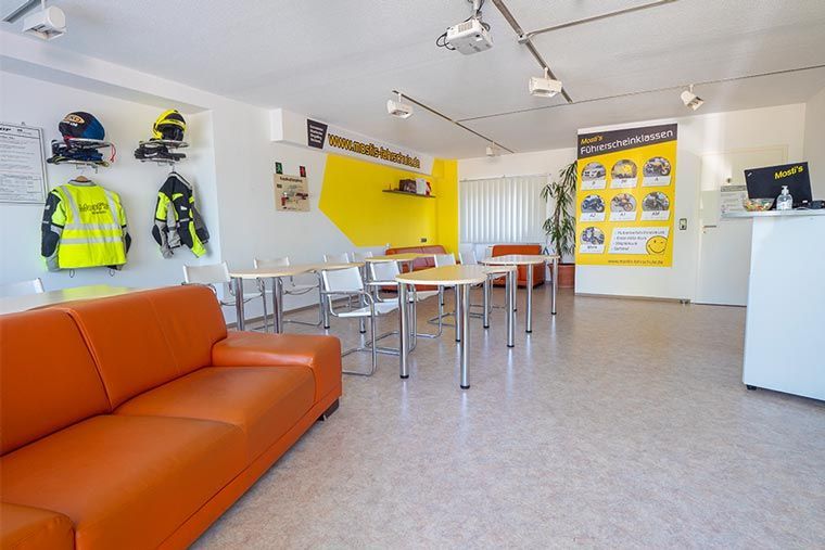 Heller Ausstellungsraum mit orangefarbenem Sofa, Tischen, Warnwesten und Helmen an weißen Wänden