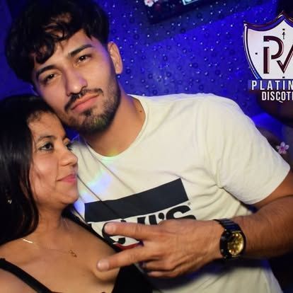 Hombre haciendo el signo de la paz con una mujer en un club; ambos sonriendo; luces del club.