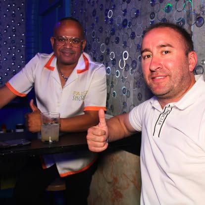 Dos hombres levantando el pulgar en un bar. Uno con camisa blanca con ribete naranja, el otro con polo blanco.