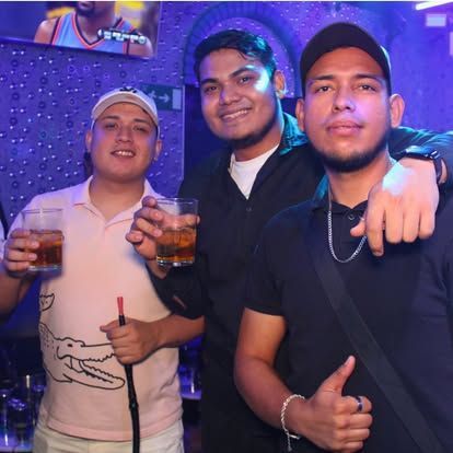 Tres hombres con bebidas en un club, sonriendo. Uno sostiene una pipa de agua iluminada con luces moradas.