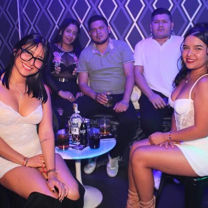 Grupo de personas en un club, sentadas alrededor de una mesa con bebidas. Iluminación tenue, gente sonriendo.