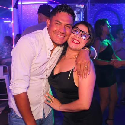 Una pareja sonriente, abrazada, en una discoteca abarrotada. El hombre con camisa blanca y la mujer con vestido negro y gafas.