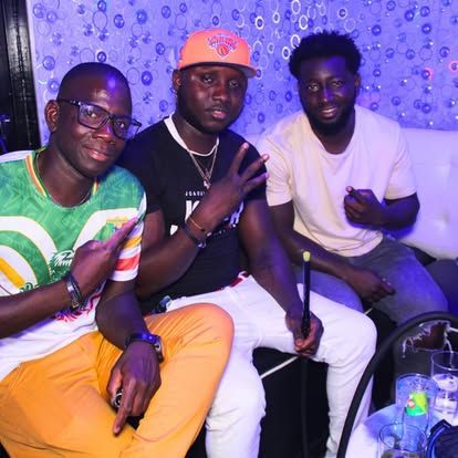 Tres hombres en un entorno poco iluminado. Uno señala, otro hace el signo de la paz y otro sostiene un objeto.