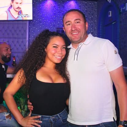 Mujer y hombre sonriendo, primer plano, en una discoteca iluminada. Ella lleva una camiseta negra y vaqueros. Él lleva un polo blanco.