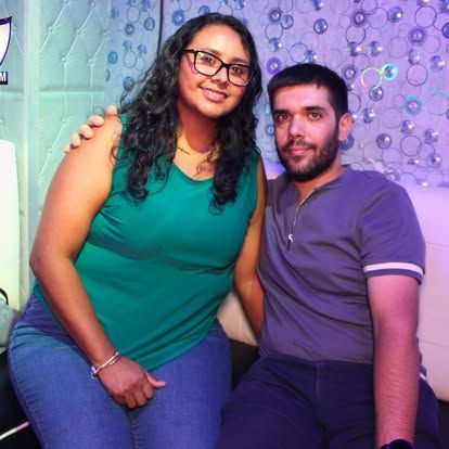 Una mujer con gafas y un hombre se sientan cerca, la mujer rodea los hombros del hombre con el brazo, sonriendo. Se sientan frente a una pared estampada.