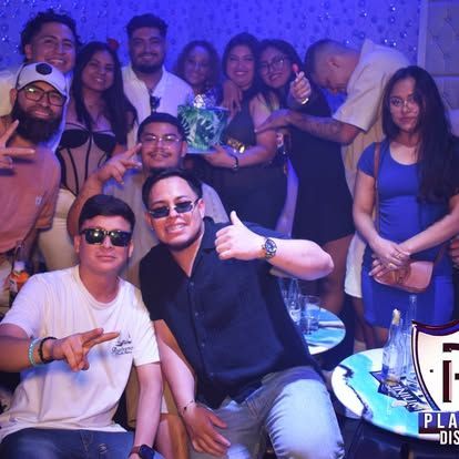 Grupo de personas posando en una discoteca, muchas sonriendo y sosteniendo una botella. Iluminación y decoración azules.