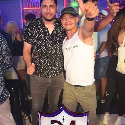 Dos hombres posan juntos en un bar lleno de gente; uno de ellos levanta el pulgar.