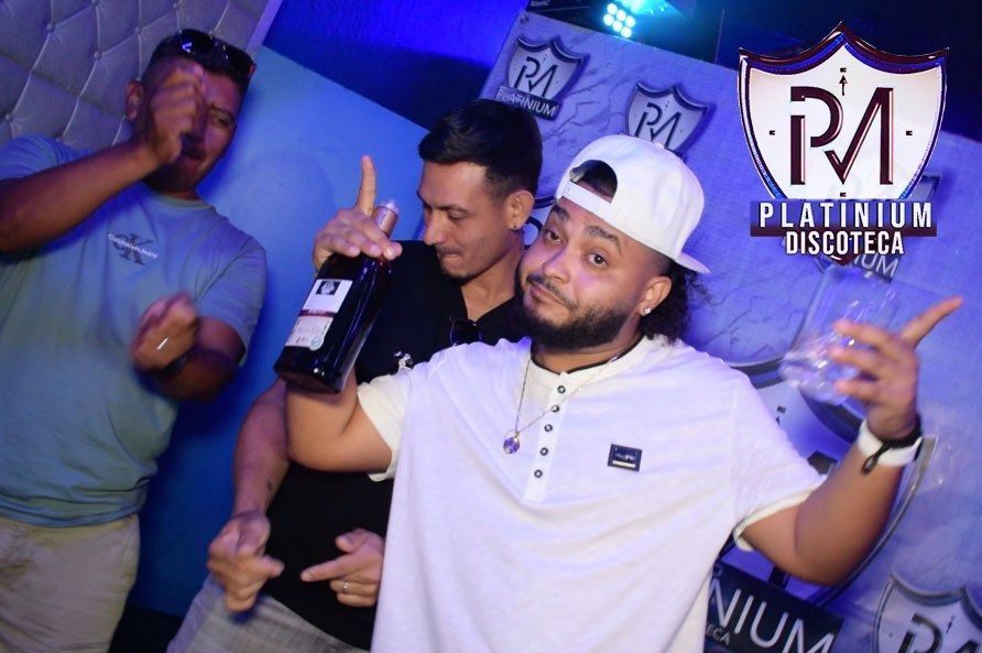 Tres hombres en la discoteca Platinium; uno con una botella en la mano y haciendo gestos, mientras los demás observan. Iluminación azul.