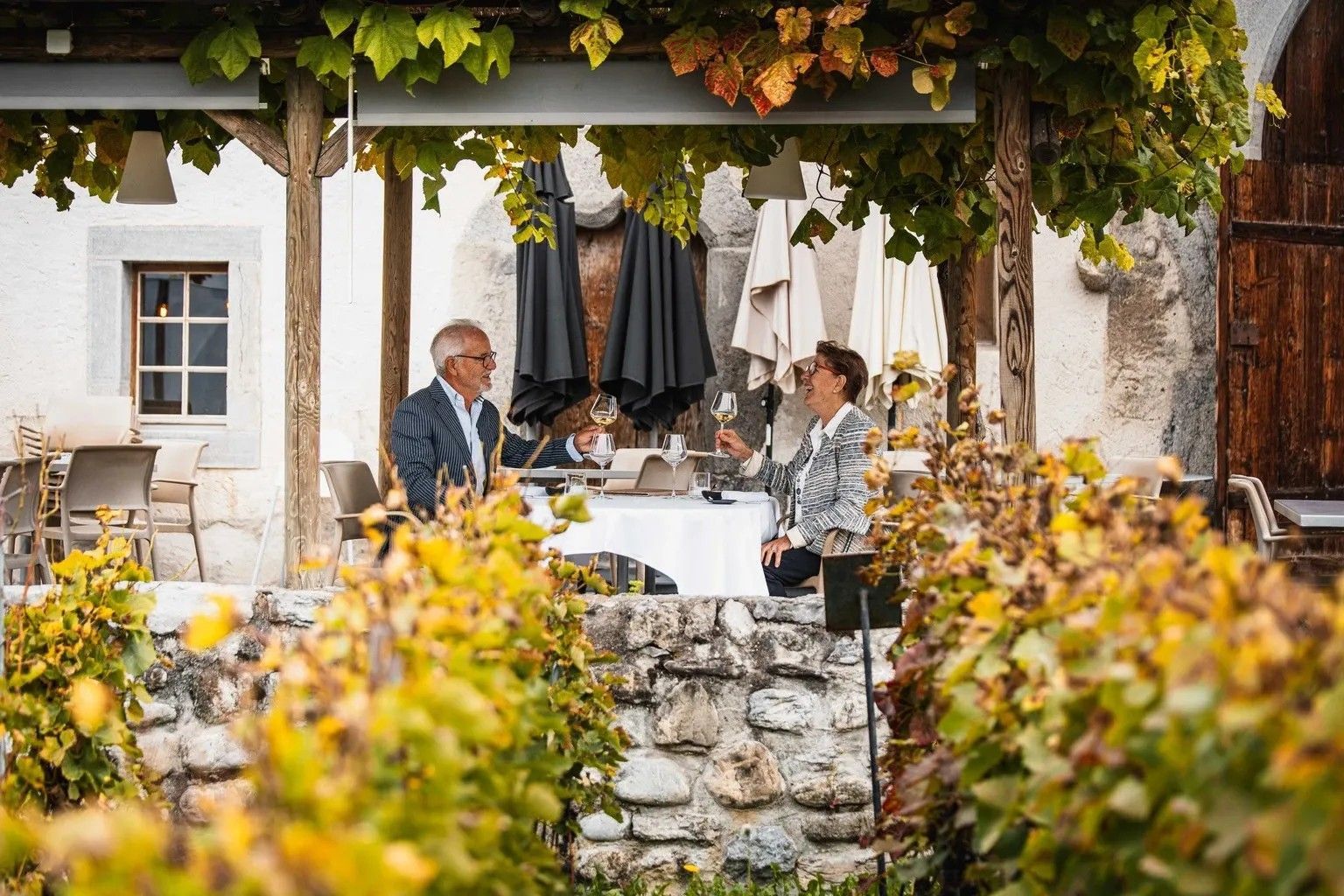 Un couple trinque au vin blanc sur la terrasse, sous la treille, entre les ceps de vigne et le mur de pierre du château